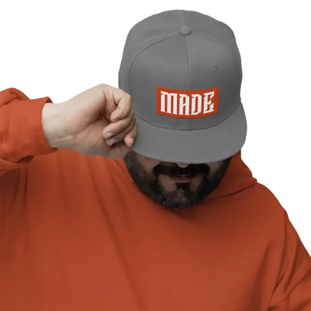Man adjusting a gray snapback hat with bold orange MADE logo on front.